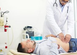 Concentrated young physician doing abdominal palpation of middle-aged patient suffering from pain, interior of modern office on background